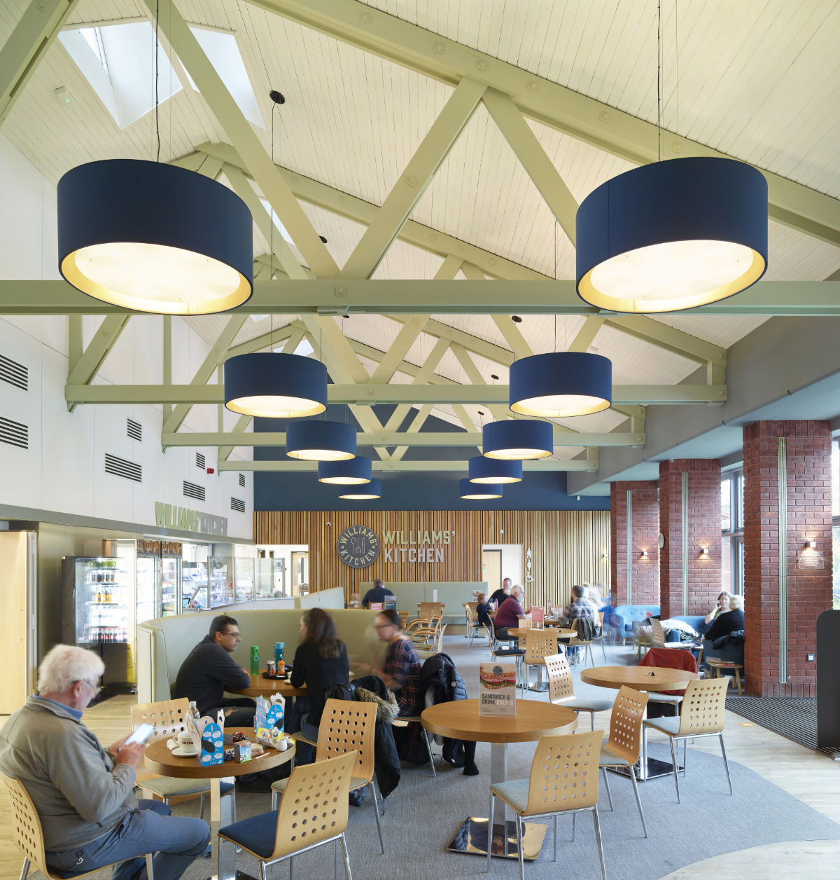Interior view of a spacious cafeteria called 'Williams' Kitchen,' featuring high ceilings with exposed trusses and large blue drum pendant lights. Customers are seated at round wooden tables and booths, enjoying food and conversation.