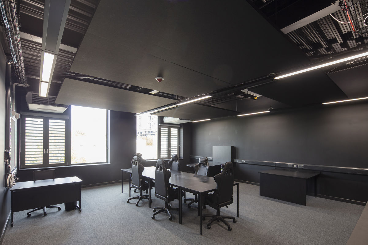 A dark-walled meeting or gaming room featuring a long central table surrounded by high-backed black gaming chairs, with a suspended black ceiling and natural light entering from windows on the left.