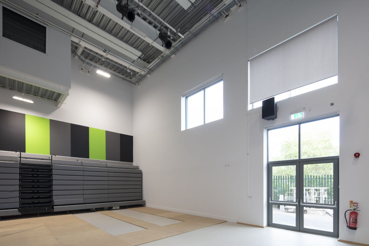 A spacious studio or performance room with high ceilings and a lighting rig. Collapsed grey bleacher seating sits against a wall decorated with black, grey, and bright green panels, next to double glass exit doors.