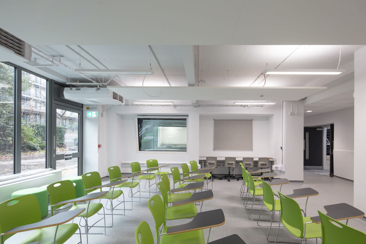 A bright classroom filled with vibrant green rolling chairs featuring attached grey writing tablets. The room has an interior observation window at the front and large exterior windows on the left.