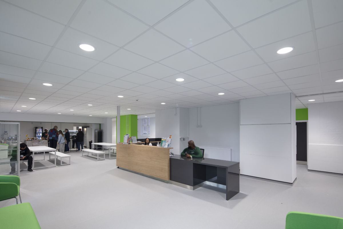 A spacious, modern lobby featuring a wood and dark grey reception desk staffed by two people. To the left is an open cafe area with several people gathering, and the space is highlighted by bright green architectural accents.