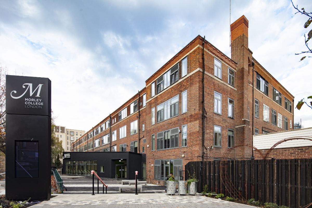 Exterior view of the Morley College London campus. A prominent black sign with the college logo stands in a paved courtyard in front of a historic, multi-story red brick building with a modern black entrance extension.