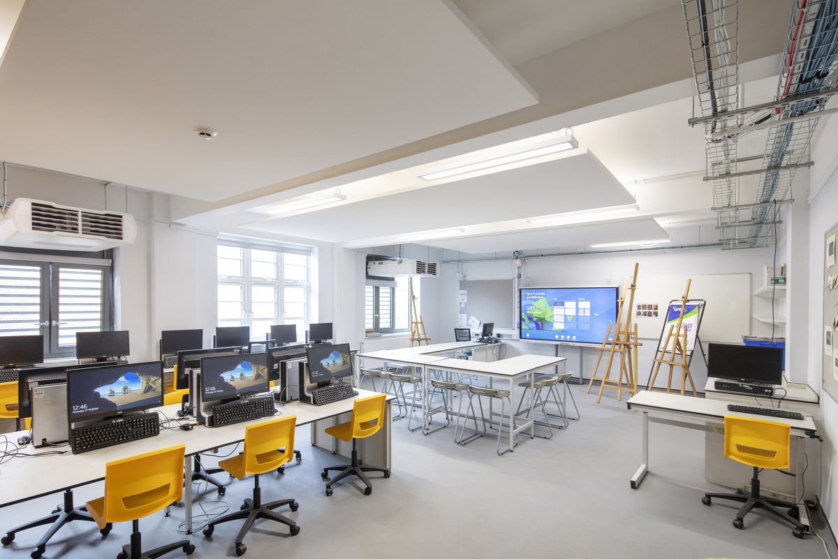 A brightly lit, modern computer lab featuring rows of desks with monitors, bright yellow rolling chairs, and a large interactive display screen next to wooden easels at the front.