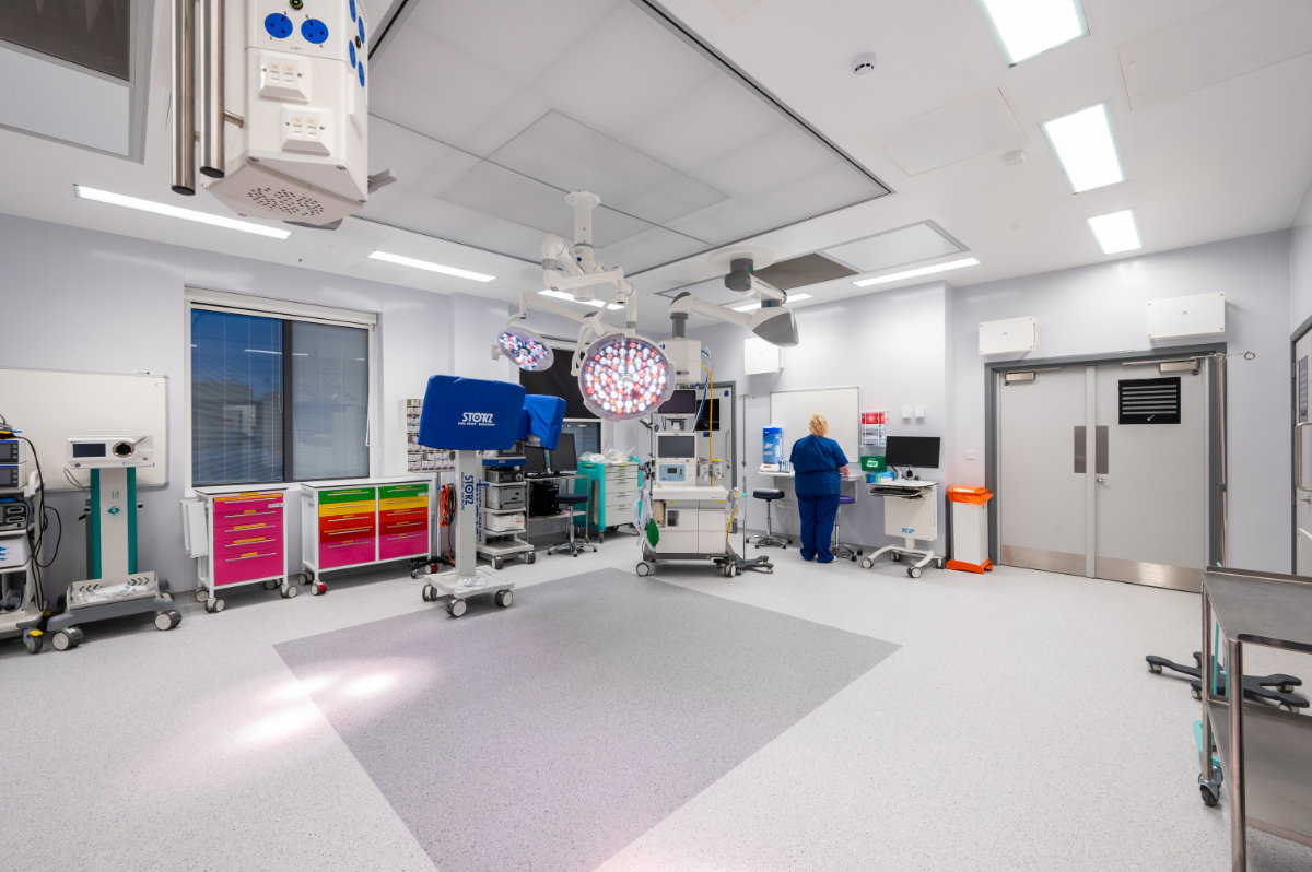 Modern hospital operating room with overhead surgical lights, anesthesia and monitoring equipment, colorful storage cabinets, and a medical staff member preparing equipment near a computer station.