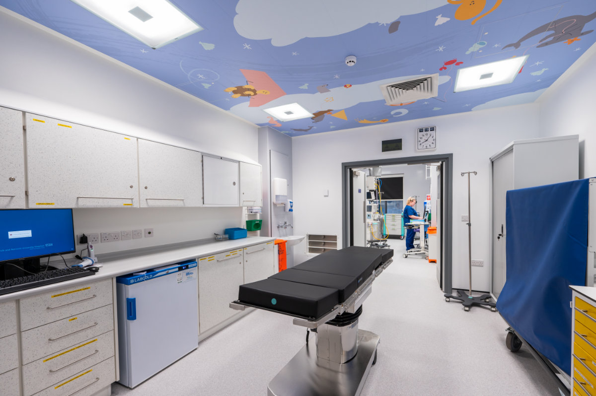 Pediatric operating room with a central surgical table, wall cabinets and computer workstation, and a ceiling painted with cartoon space-themed artwork; a medical staff member prepares equipment in the adjoining room through an open doorway.
