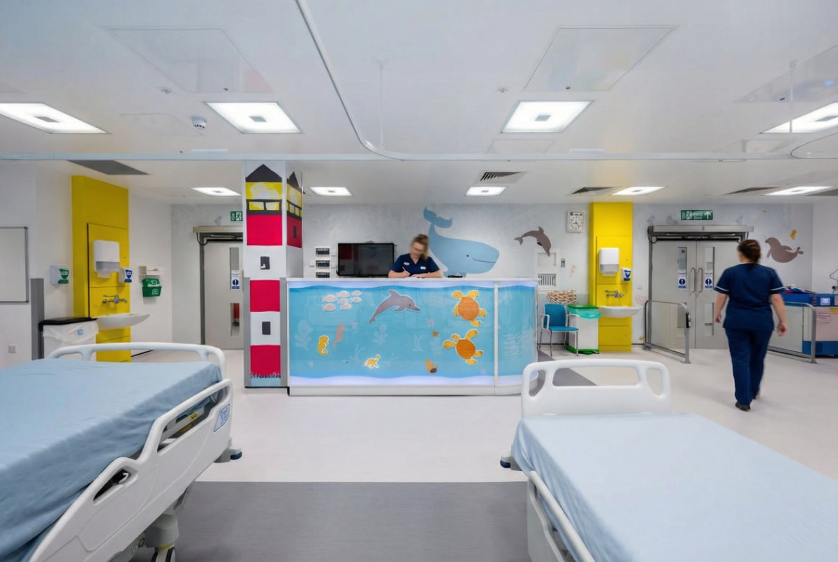 Brightly decorated pediatric hospital ward with two empty beds in the foreground and a nurse at a central station featuring colorful ocean-themed artwork, while another staff member walks toward a doorway in the background.