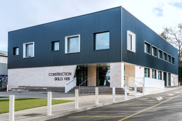 A modern two-story building labeled 'CONSTRUCTION SKILLS HUB' on the front. The structure features a mix of dark and light exterior materials, large windows, and an entrance accessed by steps. White bollards line the front, and a paved area with yellow-marked parking spaces is visible