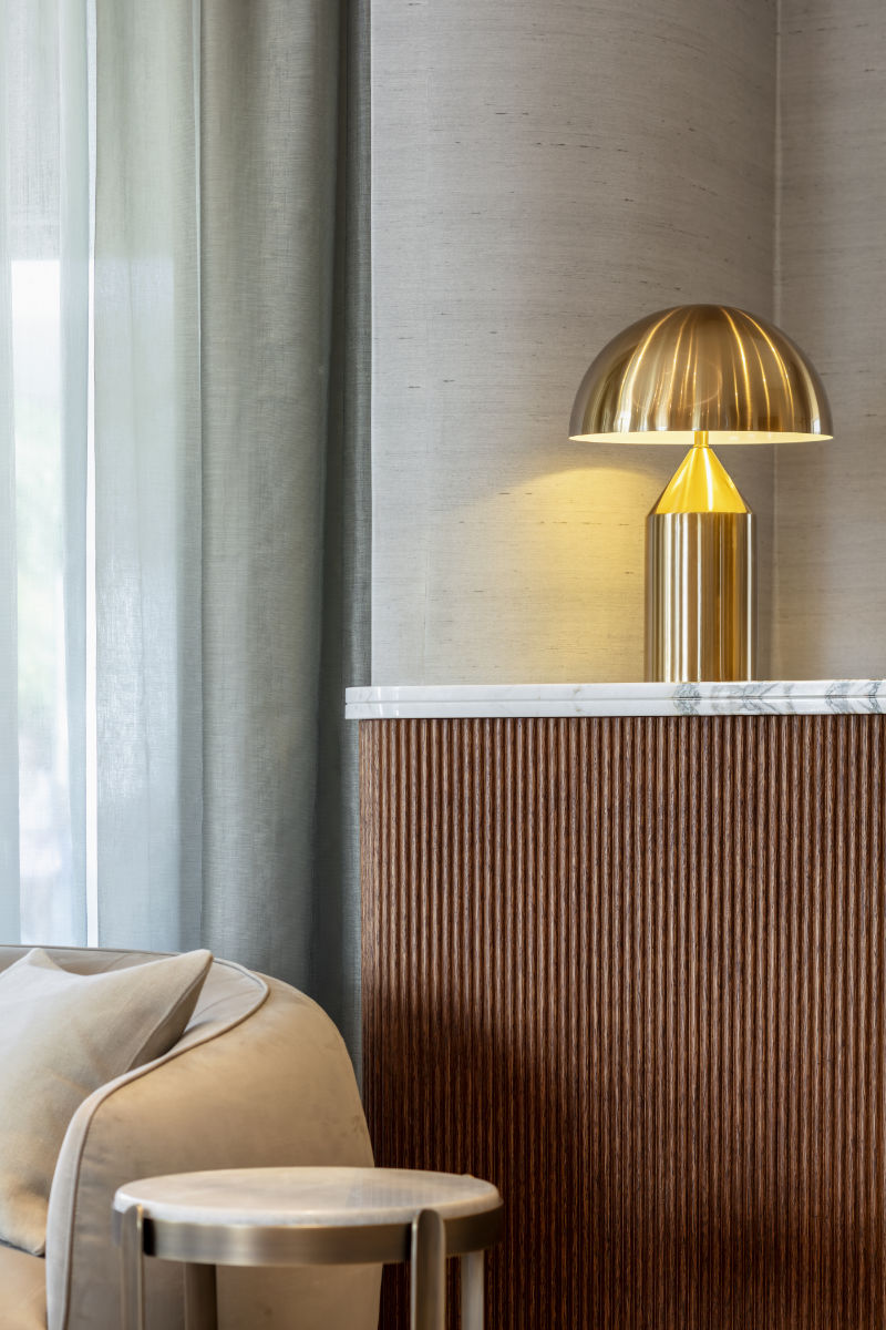 Gold table lamp with a dome-shaped shade sitting on a marble-topped wooden counter, beside sheer curtains and a beige upholstered chair in a modern interior.”