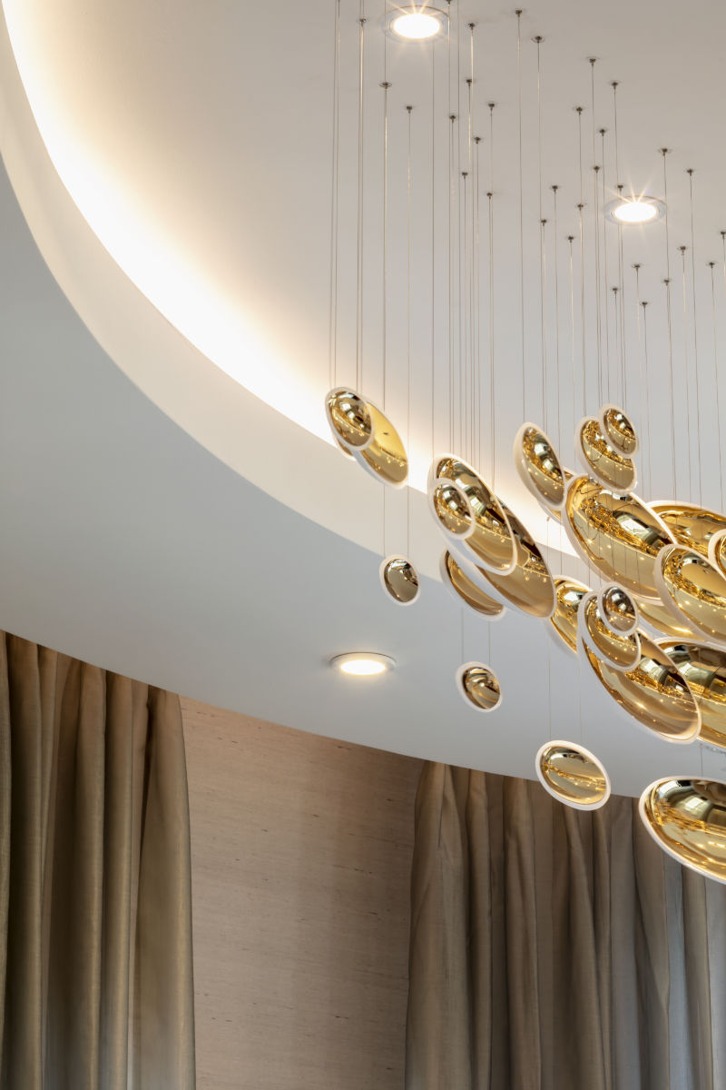 Close-up of a modern chandelier with multiple gold, oval-shaped pendants suspended from thin wires beneath a curved white ceiling, with soft ambient lighting and curtains in the background.