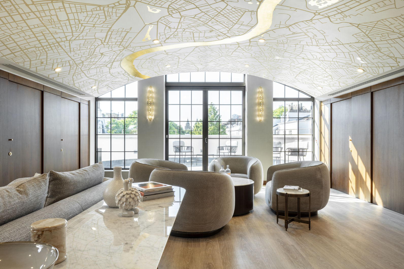 Elegant lounge with curved upholstered seating, marble-topped tables, and a decorative ceiling patterned like a city map, opening onto a balcony through large glass doors with daylight streaming in