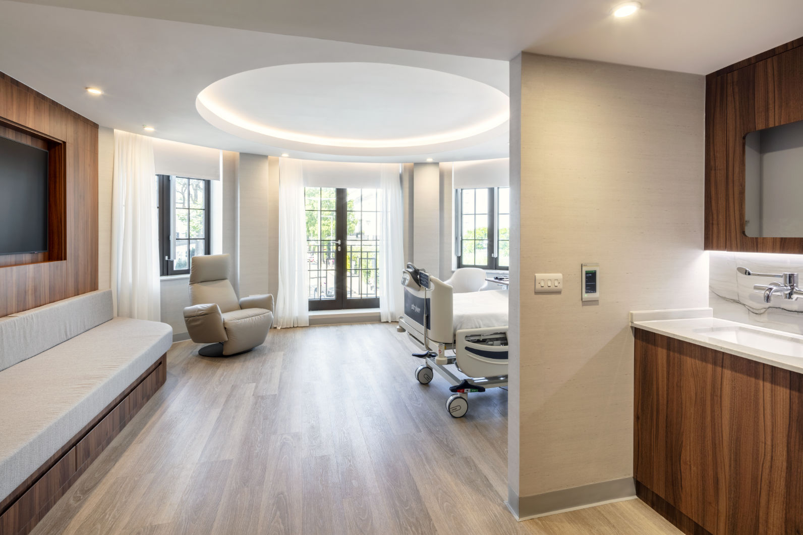 Modern hospital room with a patient bed, recliner chair, built-in bench with a TV, large windows, and a wooden vanity area.