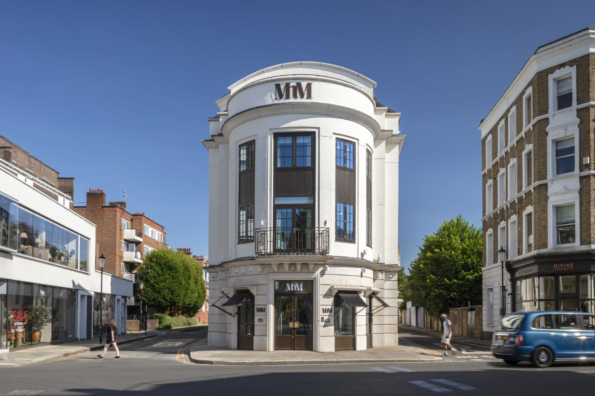 Street view of a distinctive white, curved corner building with large windows and ‘MM’ signage, set at an intersection with nearby shops, pedestrians walking by, and a blue car passing under a clear blue sky