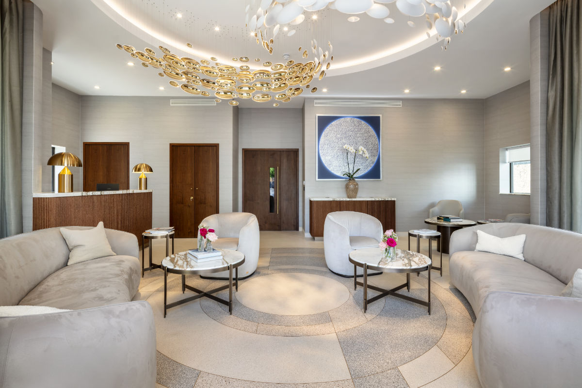 Elegant reception area with curved light grey sofas, cream armchairs, marble-topped coffee tables, and a dark wood reception desk with gold lamps. A large, sculptural chandelier hangs from the ceiling, and a framed artwork featuring white orchids is displayed on the wall.